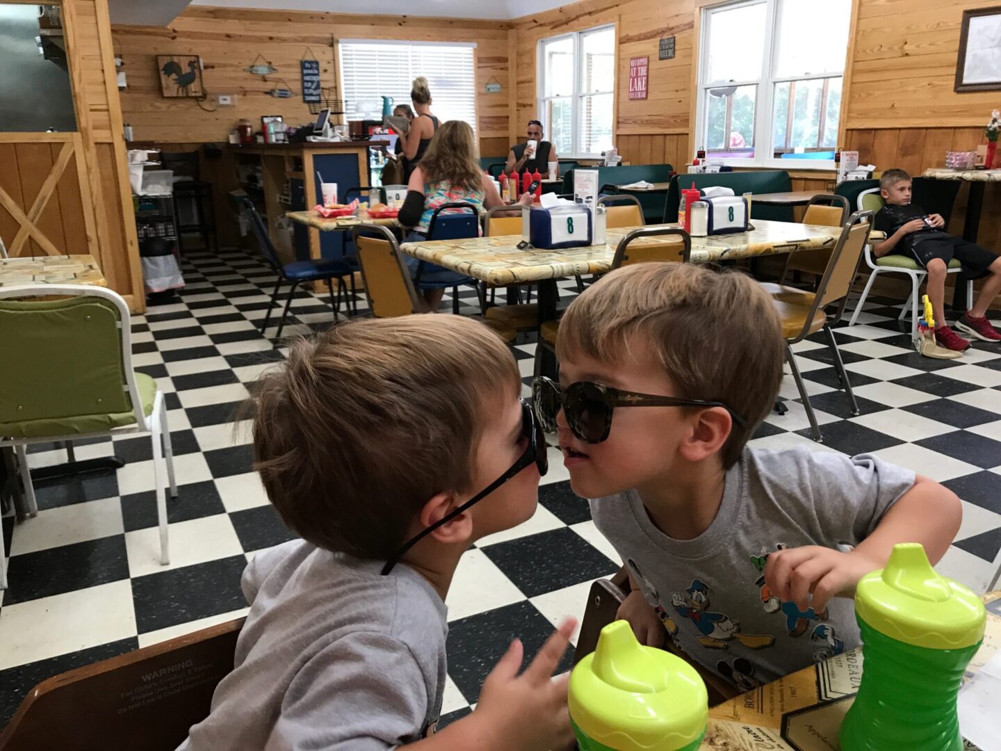 Two young boys are sitting at a table and one is kissing the other.